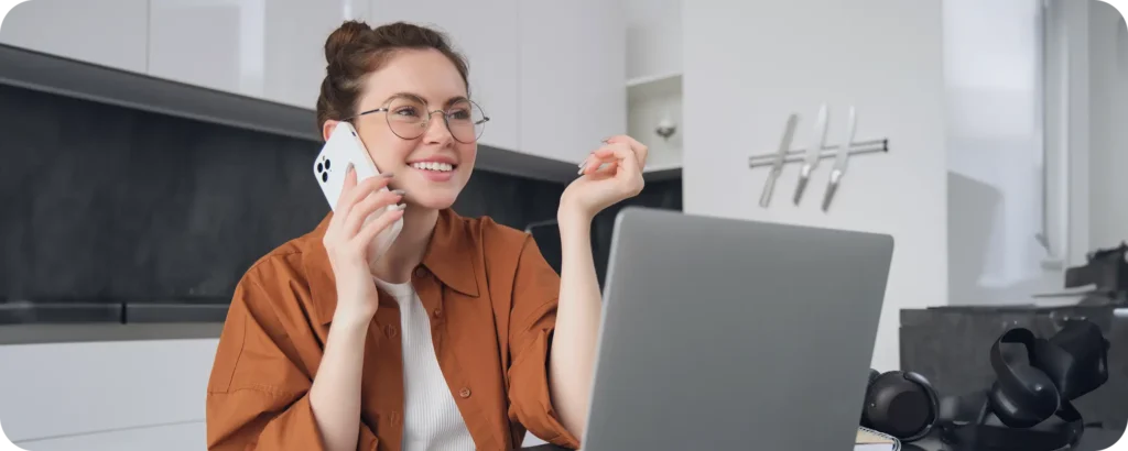 Mulher sorridente em ligação por celular, sentada em frente ao notebook — representando o atendimento próximo, acessível e acolhedor oferecido pela equipe da Volare.