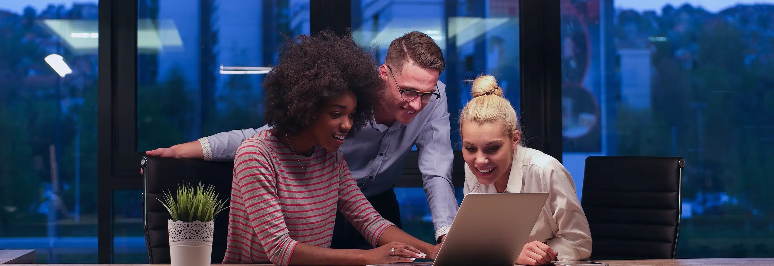 Três profissionais sorrindo enquanto trabalham juntos em um notebook, em ambiente corporativo noturno — representando colaboração, criatividade e engajamento em equipe, valores cultivados pela Volare.